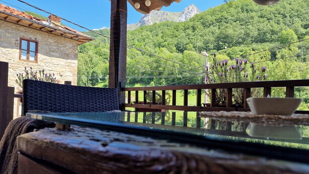 Una mesa de madera rústica con cubierta de vidrio, una silla de mimbre y vistas a un paisaje verde exuberante con montañas al fondo.