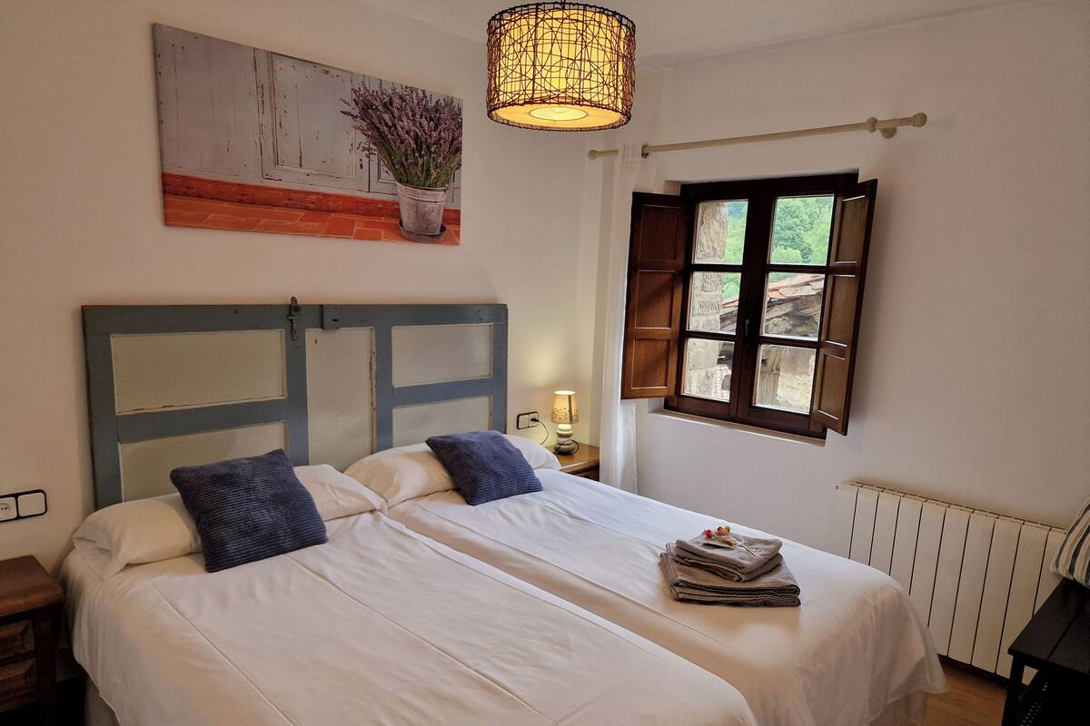 A double bed with white linens and two pillows, a wooden headboard, a window with wooden shutters, a hanging light fixture, and a wall-mounted painting of lavender.