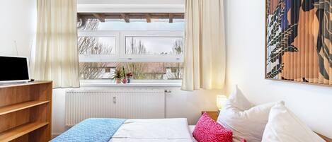 A bedroom with a bed, a desk, a window with curtains, a radiator, and a painting on the wall.