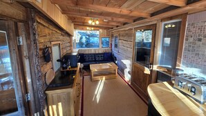 Interior rústico de una cabaña con techo de madera, kitchenette, sofá de cuero negro y un ventanal con vistas a un bosque.