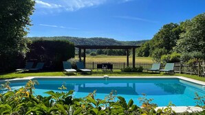 A swimming pool with lounge chairs and a pergola, surrounded by greenery and a view of hills. - The Farmhouse (MEYRONNE)