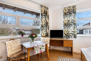 Huoneessa on puinen työpöytä, tuoli, sänky, televisio ja ikkuna, jossa on verhot.