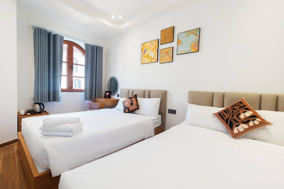 A hotel room with two beds, a wooden nightstand, a desk, a chair, and a window with curtains.