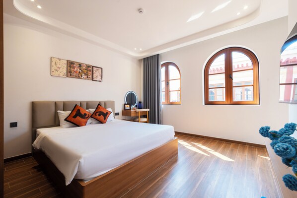 Una habitación de hotel moderna con una cama grande, suelo de madera, un tocador con espejo y dos ventanas de arco.