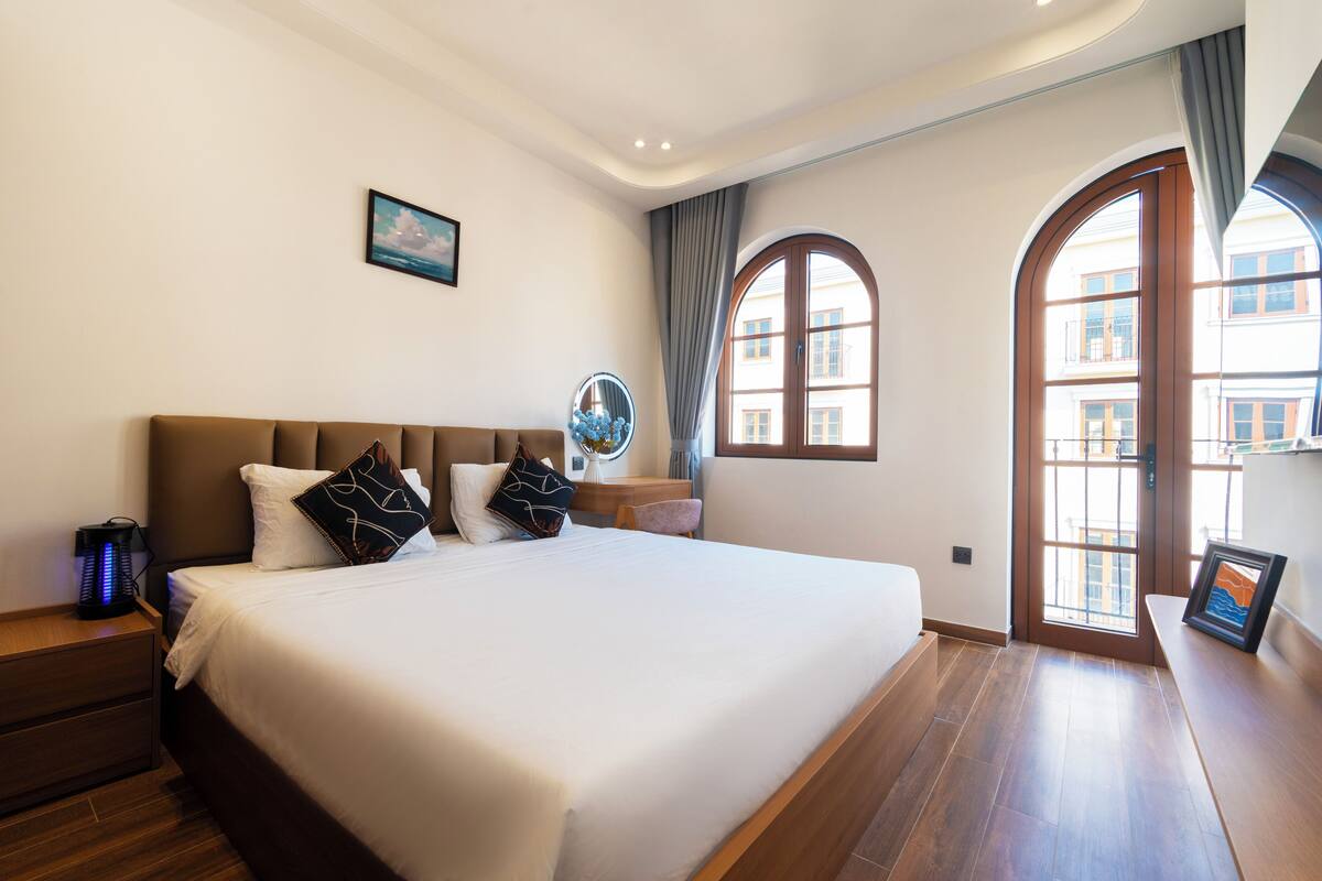 A hotel room with a large bed, a nightstand, a desk, and a view through a window.