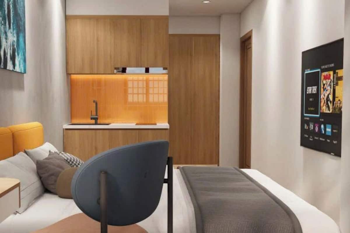 A modern hotel room with a bed, a desk, a chair, a small kitchenette, and a wall-mounted TV.