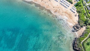 Vista aerea di una spiaggia con acque cristalline turchesi, una battigia sabbiosa e una fila di ombrelloni.