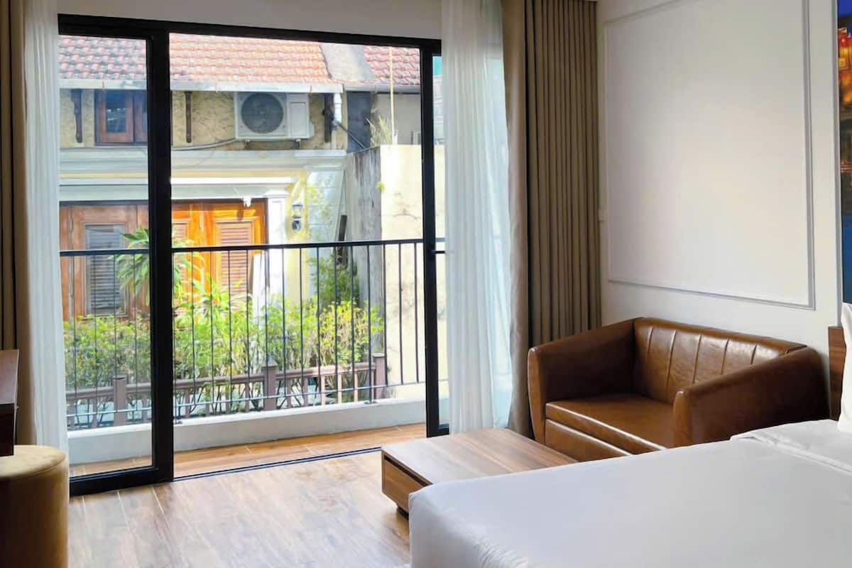 Een hotelkamer met een groot bed, een bank, een houten vloer en een balkon met uitzicht op gebouwen en groen.