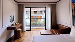 Une chambre d’hôtel avec un grand lit, un plancher en bois, un bureau avec un miroir, un canapé en cuir marron et un balcon offrant une vue sur les bâtiments et la verdure.