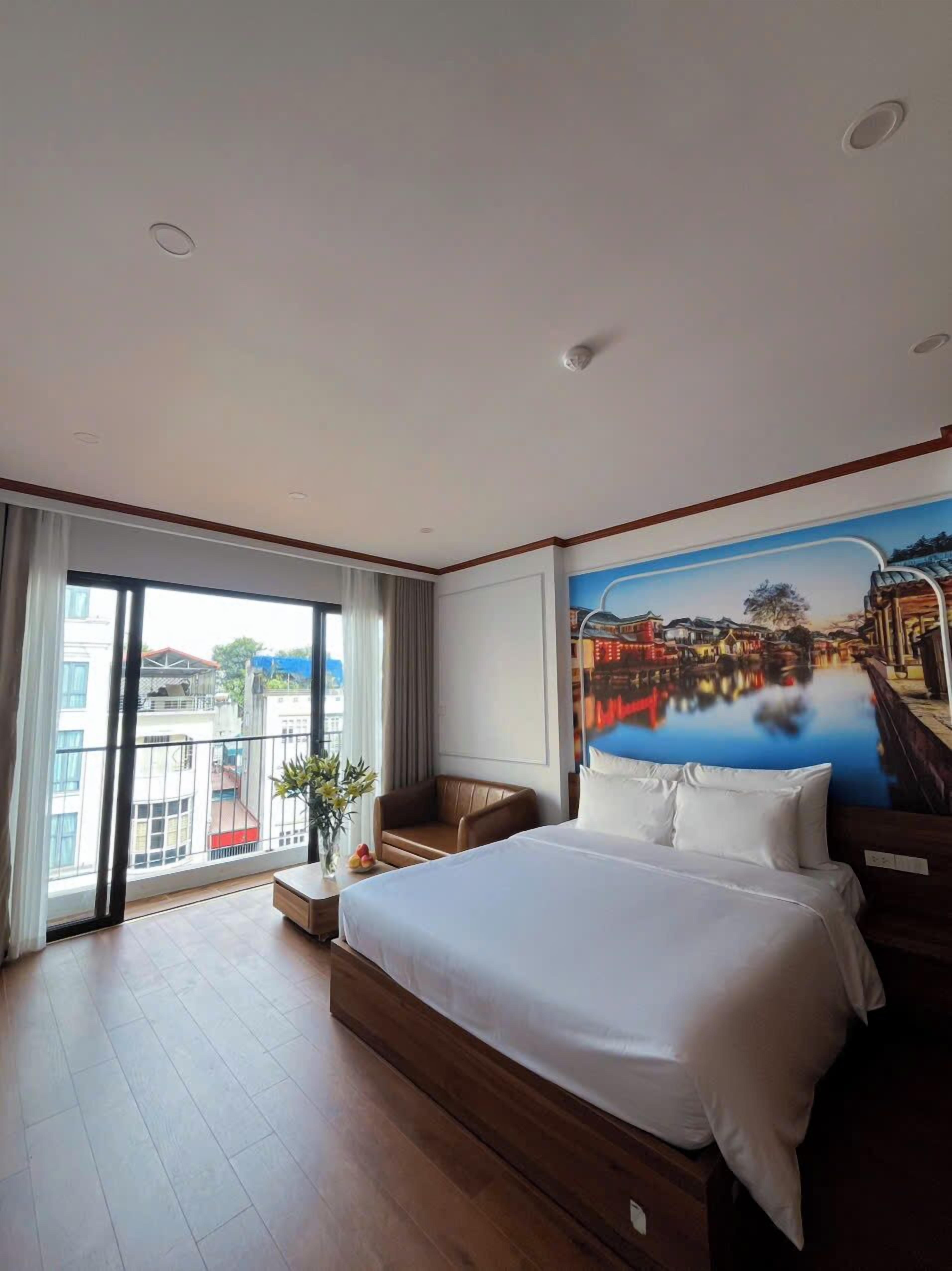Een hotelkamer met een groot bed, uitzicht op de stad vanaf een balkon en een muurschildering van een rivierlandschap.