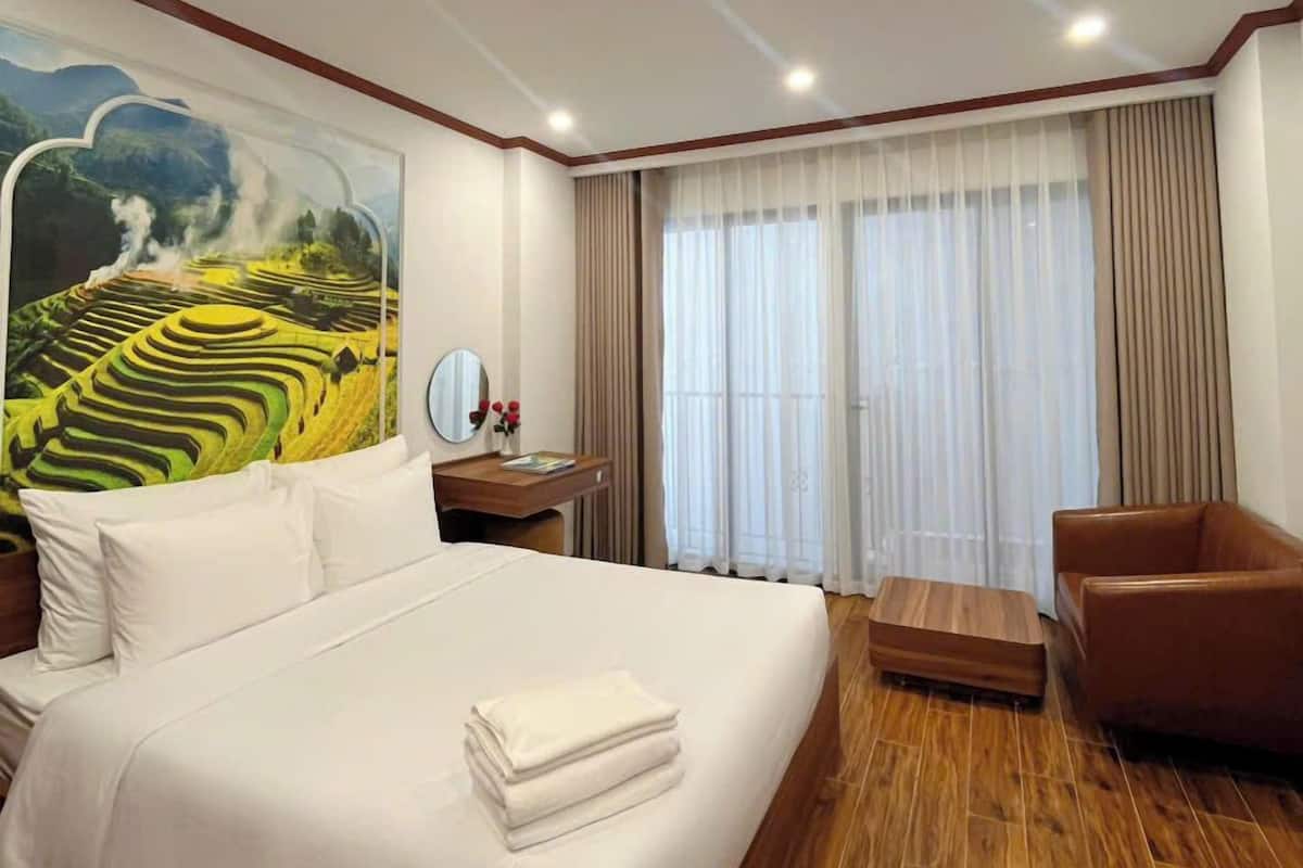 Een hotelkamer met een groot bed, een houten vloer, een bureau met stoel, een raam met gordijnen en een schilderij aan de muur.
