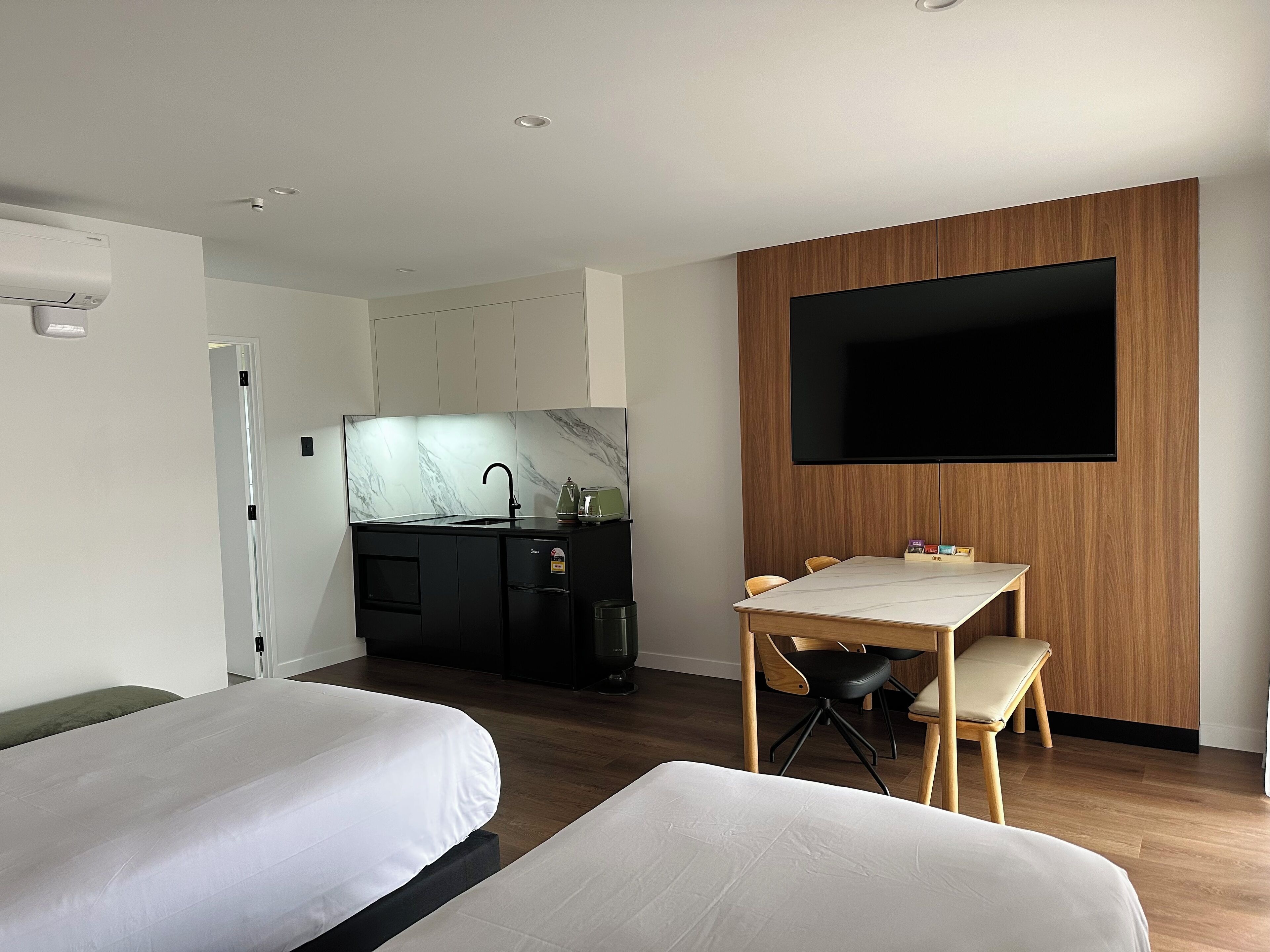 A modern hotel room with two beds, a kitchen area, a dining table with chairs, and a flat-screen TV mounted on the wall.