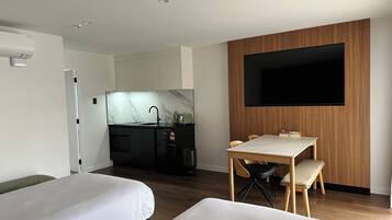 A modern hotel room with two beds, a kitchen area, a dining table with chairs, and a flat-screen TV mounted on the wall.