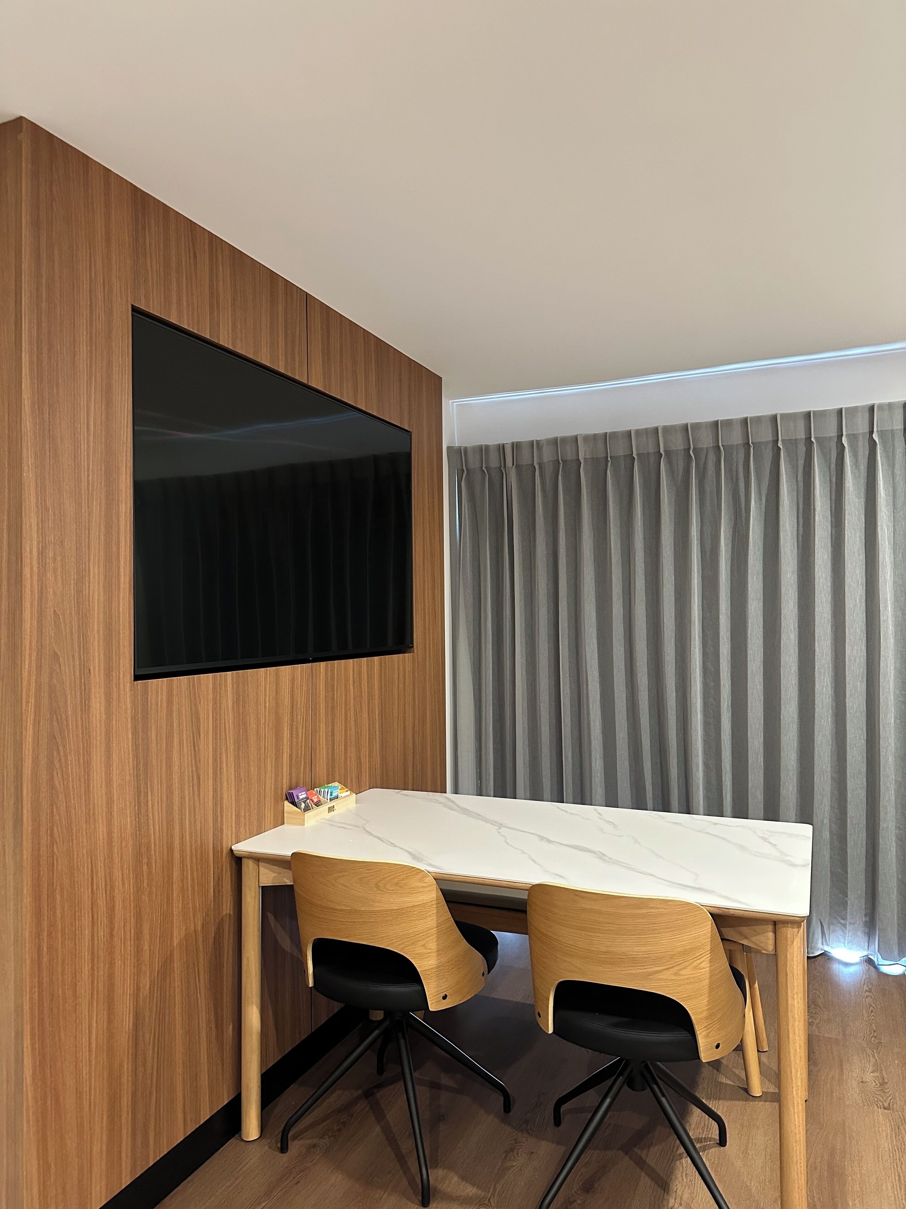 Una habitación moderna con pared de madera, un televisor de pantalla plana grande, una mesa de mármol blanco, dos sillas de oficina negras y una cortina gris.
