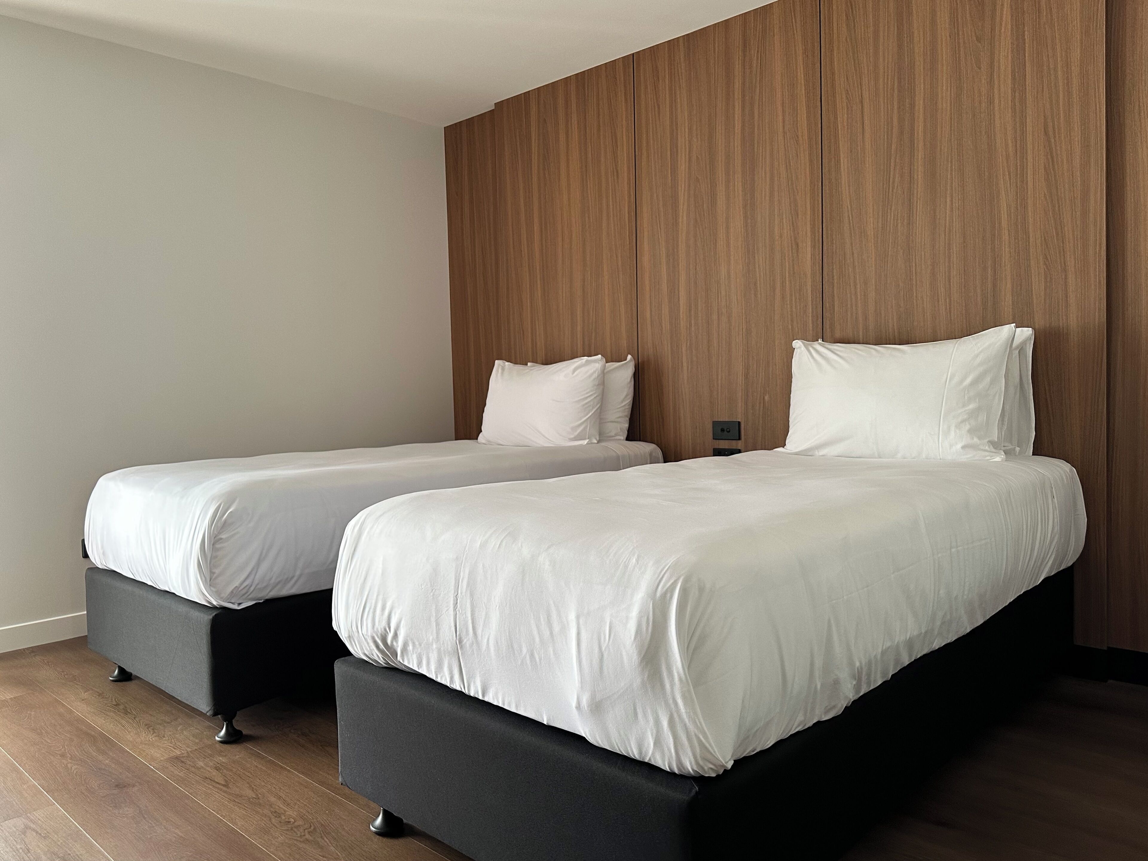 A modern hotel room with two beds, wooden paneling on the wall, and a wooden floor.