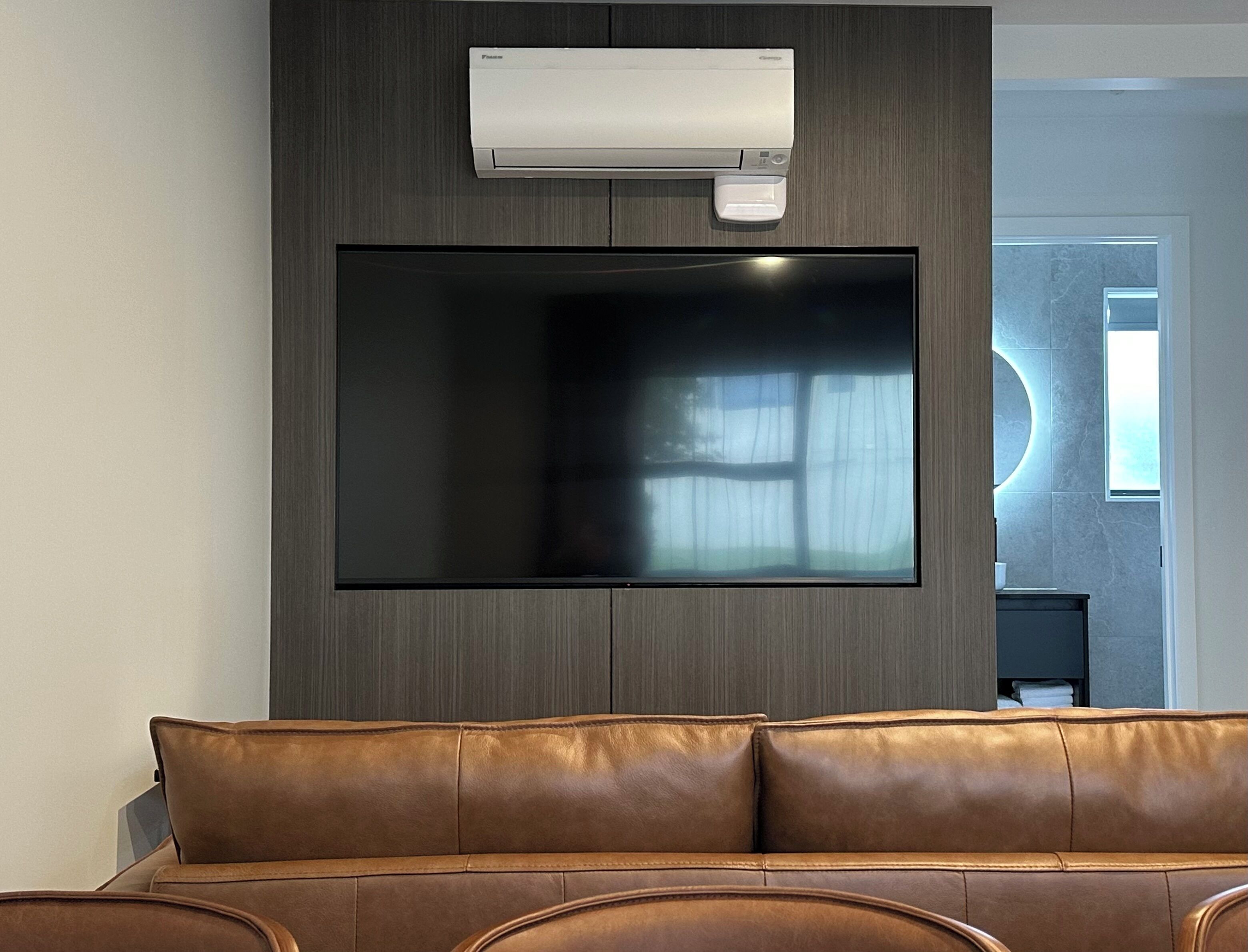 A modern living room with a large flat-screen TV mounted on a dark wood wall, a leather sofa, and a built-in air conditioning unit.