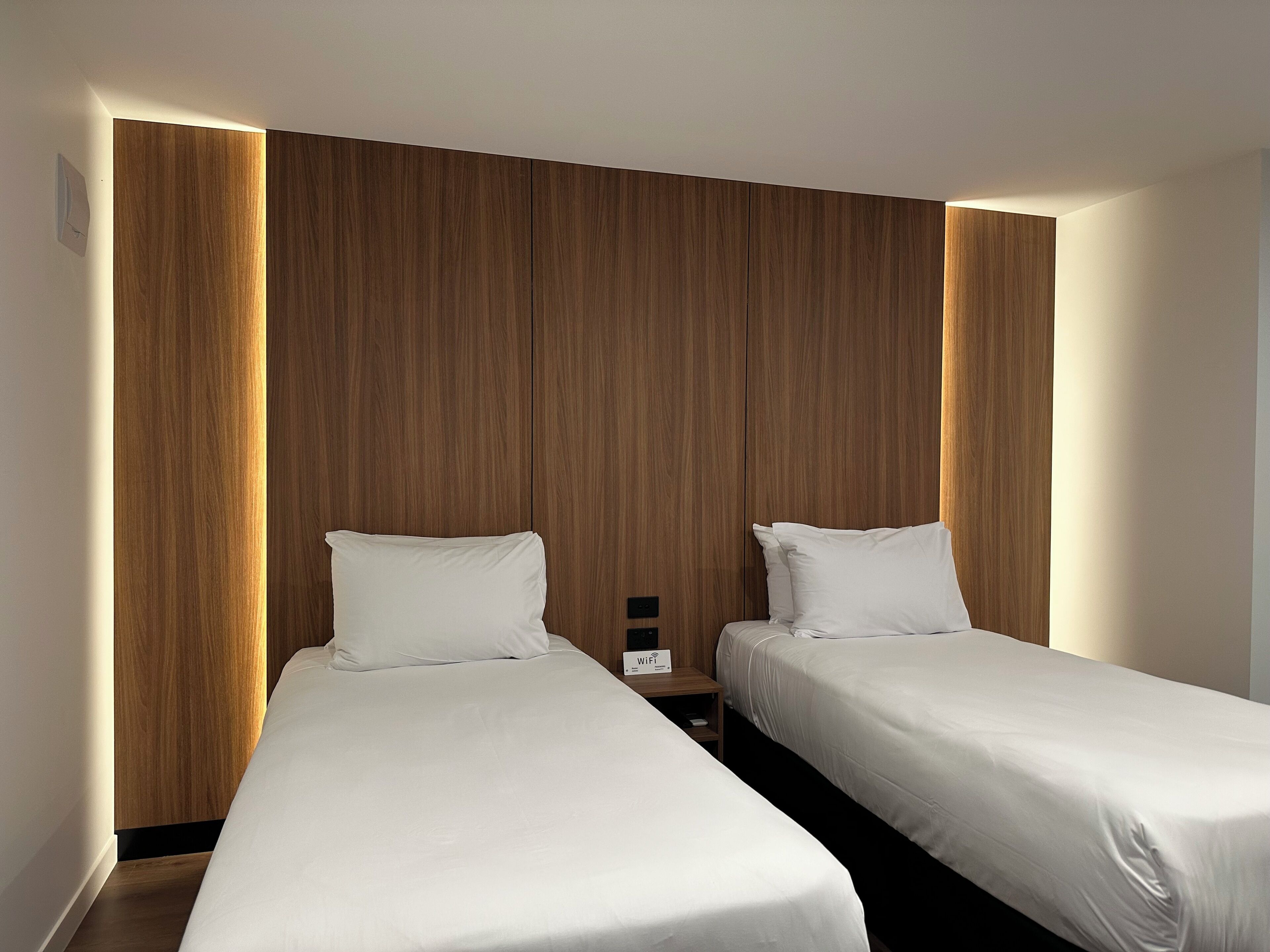 A hotel room with two single beds, wooden paneling on the walls, and a nightstand with a Wi-Fi sign.