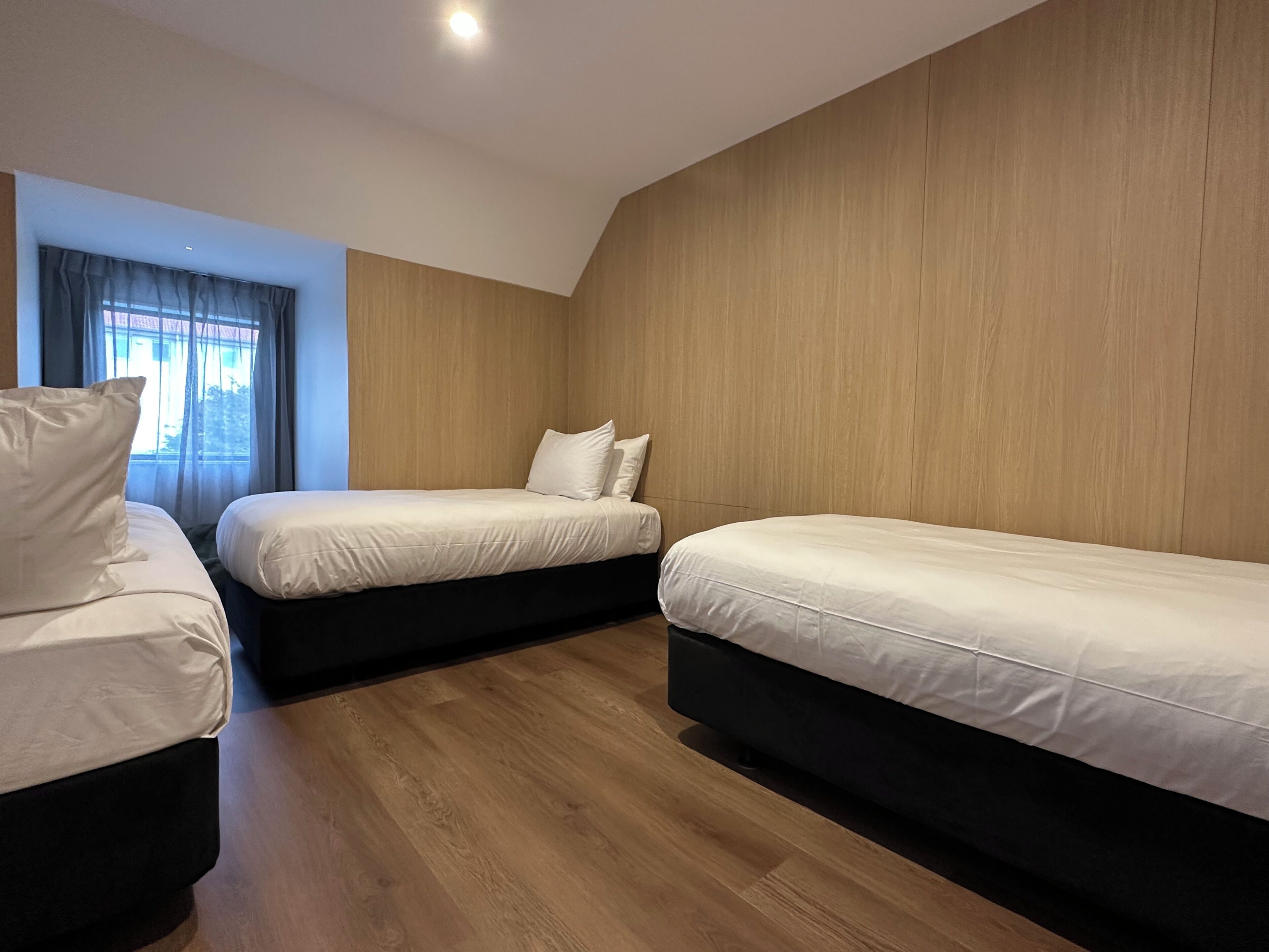 A hotel room with two single beds, a wooden floor, and a window with curtains.