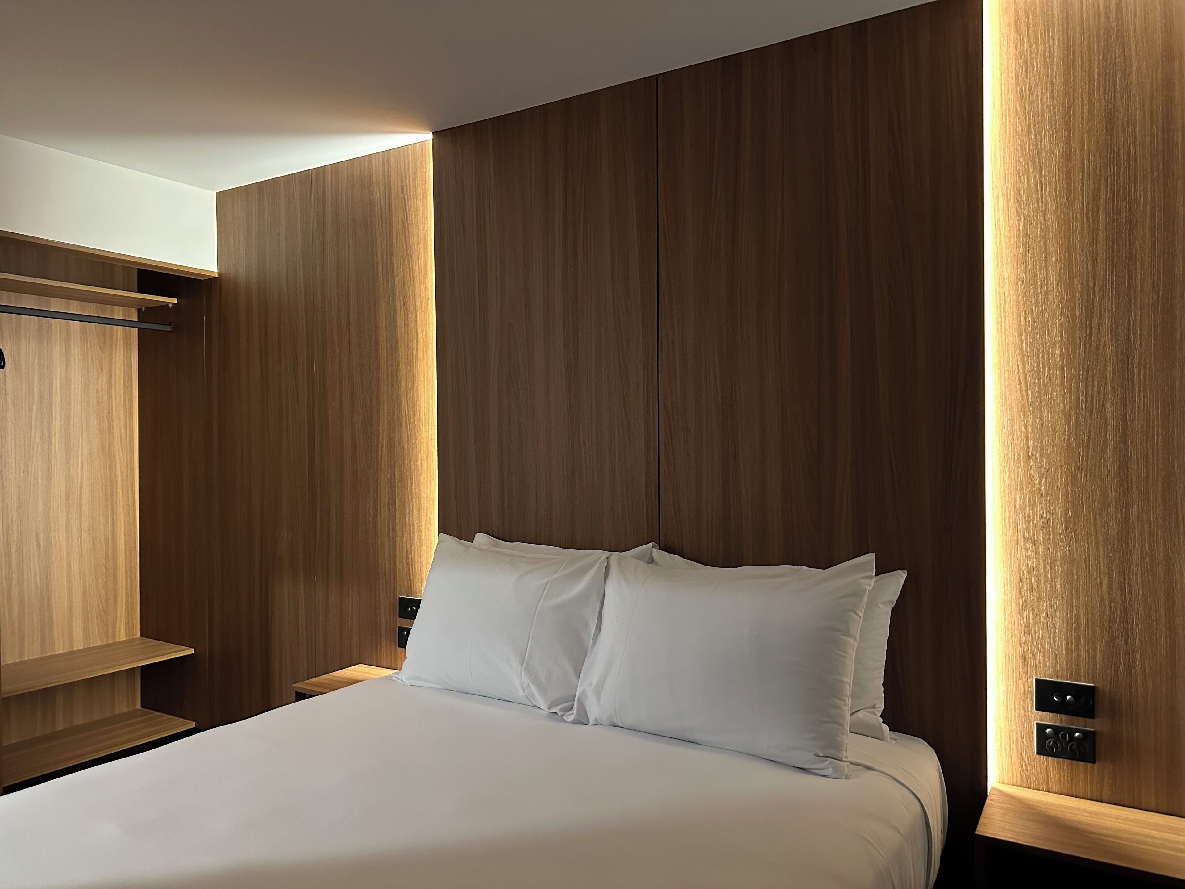 Una habitación de hotel moderna con un cabecero de madera, una cama bien tendida y una estantería empotrada.