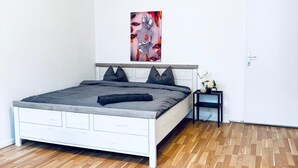 Ein Schlafzimmer mit Holzboden, einem weißen Bett mit grauer Bettwäsche, einem kleinen Nachttisch mit Blumenvase und einer Tür.