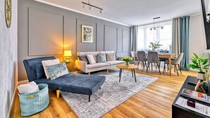 A modern living room with a sofa, armchair, and coffee table, a dining area with chairs, and a large window with curtains. - Apartamenty Sun & Snow Pod Ratuszem (Jelenia Góra)