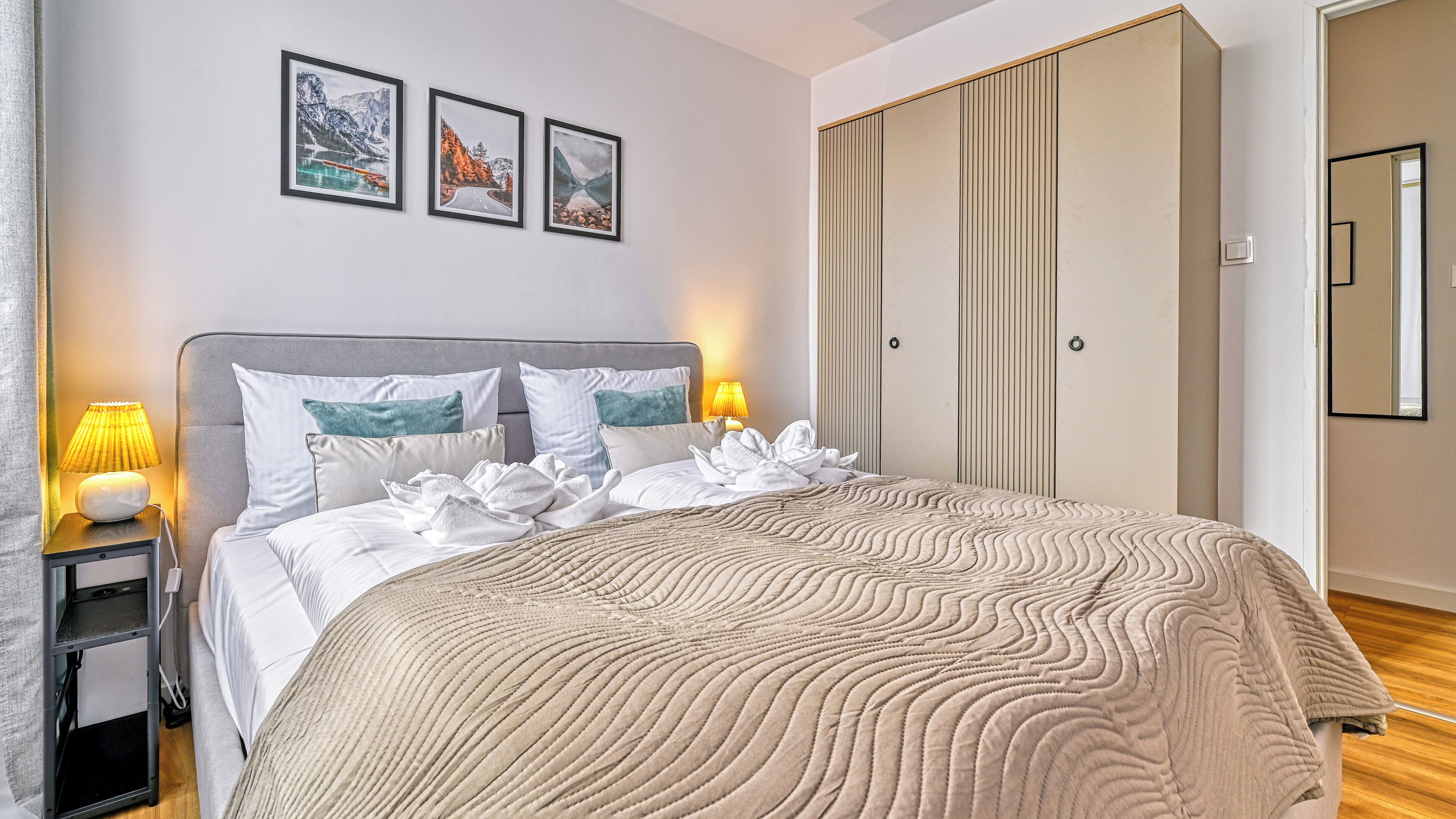 Un lit bien fait, recouvert d’une couverture à motifs, une lampe de chevet et une table de nuit. Trois tableaux encadrés sont accrochés au mur au-dessus du lit.