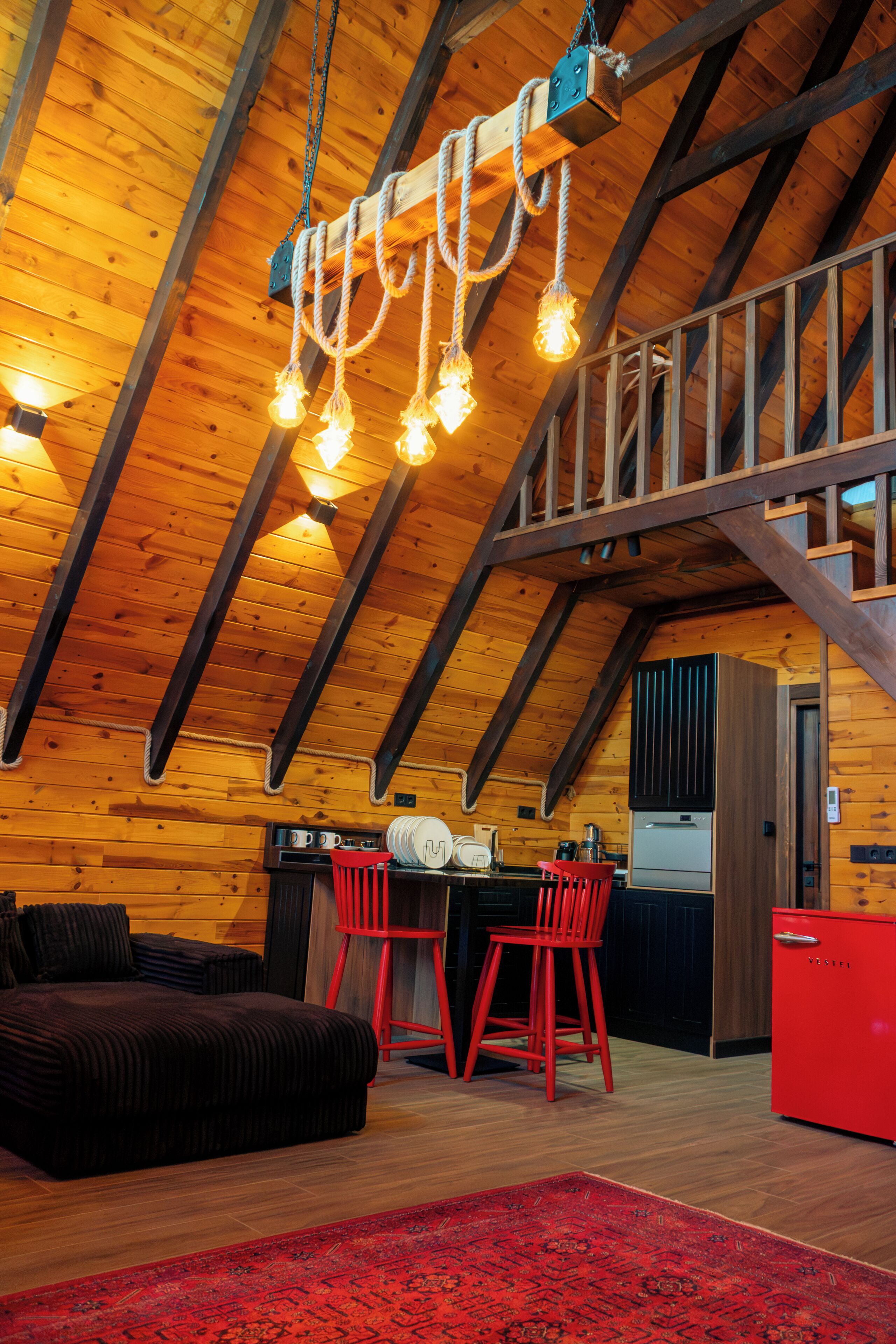 A cozy log cabin interior with a loft bedroom, a kitchen with a red refrigerator, and a dining area with red chairs.