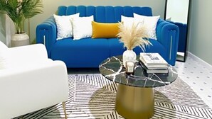 A living room with a blue sofa, white armchair, and a black and gold coffee table. There is a painting on the wall and a floor lamp. - Naivasha Budget Suites  (Naivasha)