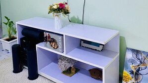 A living room with a flat-screen TV mounted on the wall displaying a fireplace image. Below the TV, there is a white entertainment unit with shelves, a small potted plant, and a vase with flowers. - Naivasha Budget Suites  (Naivasha)