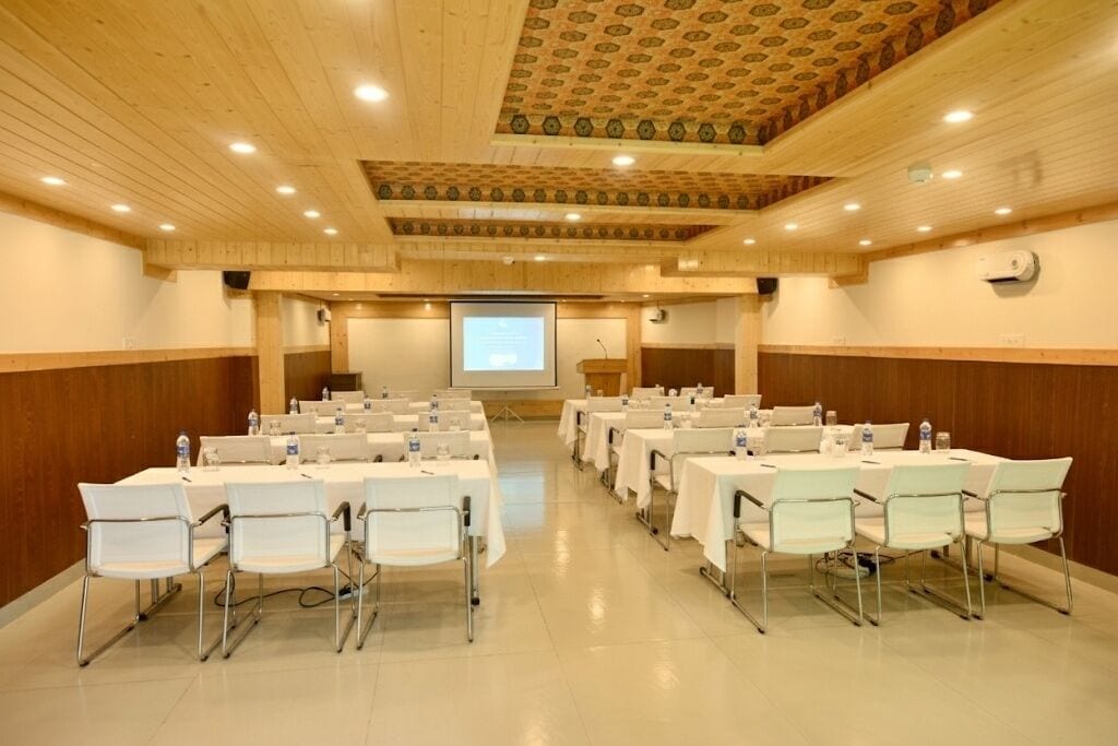 Un salón de conferencias con mesas y sillas blancas, una pantalla de proyector y paneles de madera.