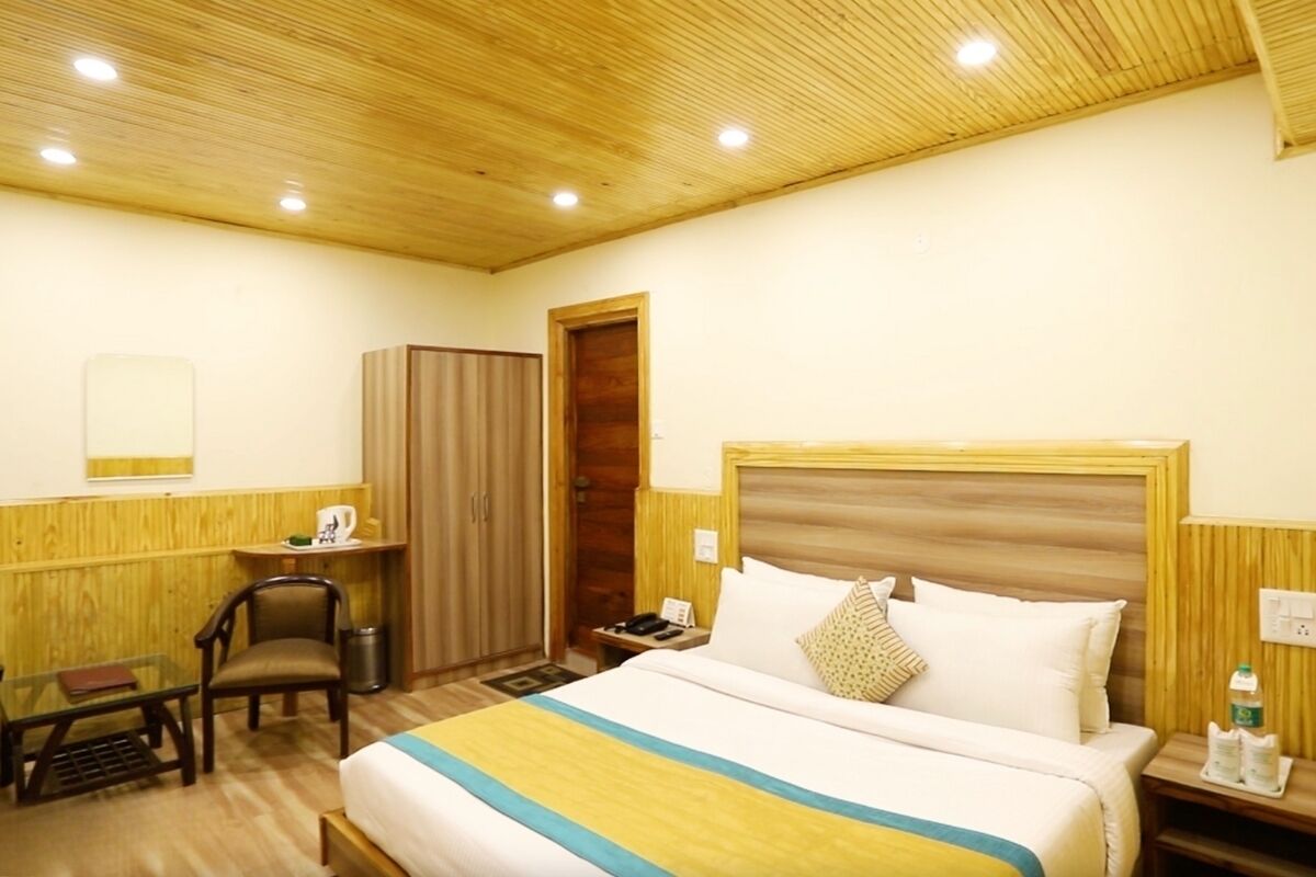 A hotel room with a wooden ceiling, a bed with a striped bedspread, a desk with a chair, a wardrobe, and a nightstand with a phone and a bottle.