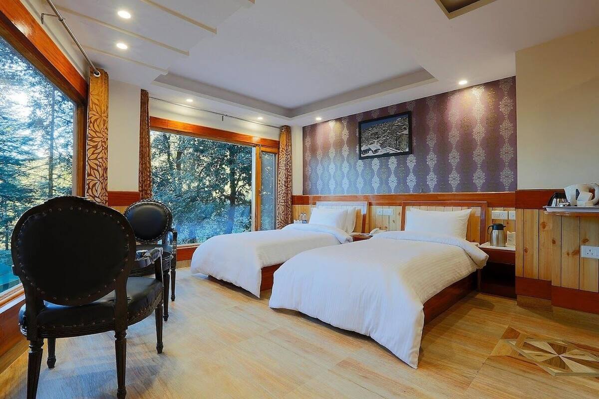 A hotel room with two beds, a wooden chair, a table, and a view of trees through large windows.