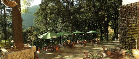 An outdoor patio area with tables and chairs, surrounded by trees and potted plants.
