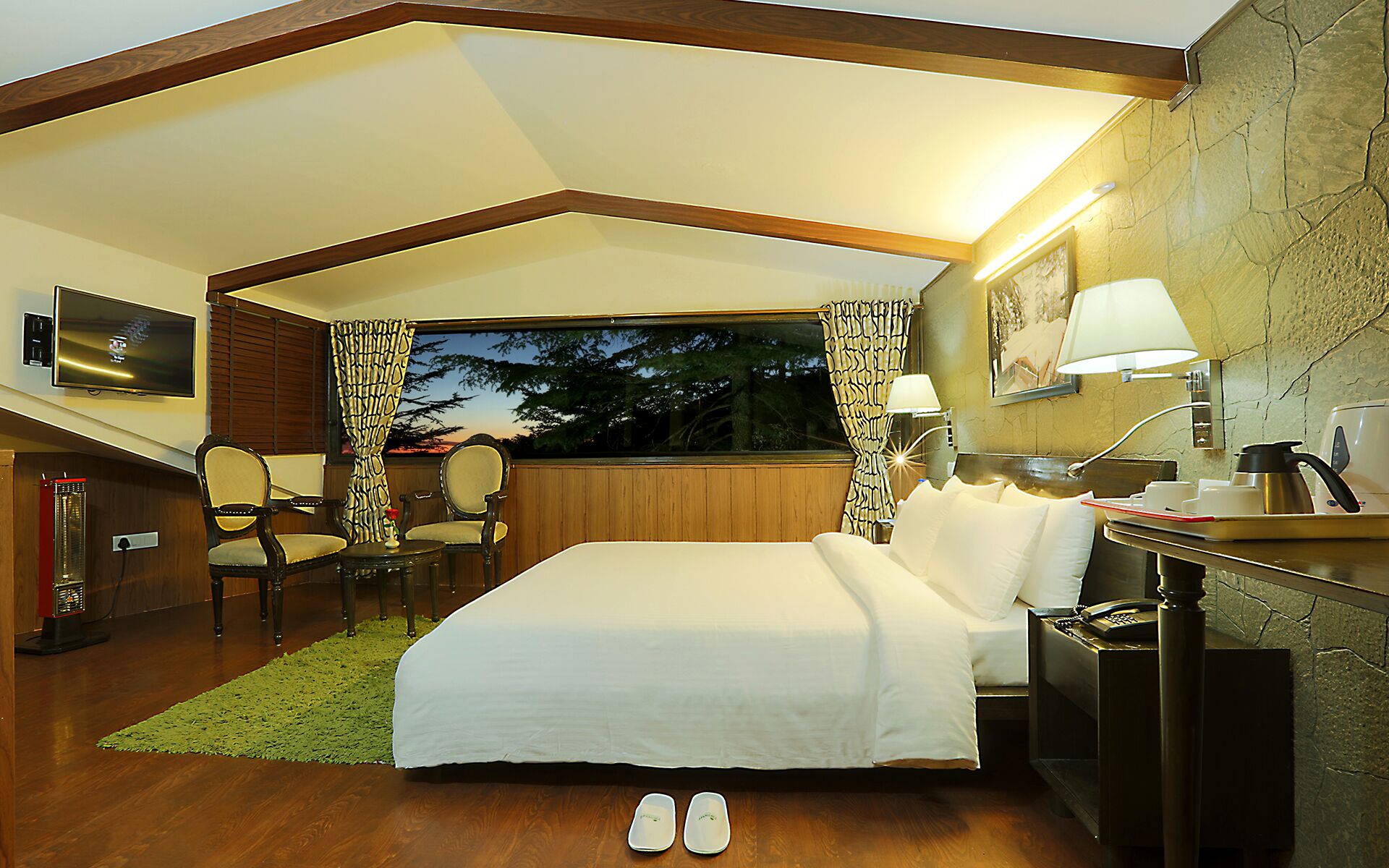 A hotel room with a bed, two chairs, a desk with a lamp, a television, and a window with curtains.