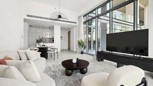A modern living room with a sofa, coffee table, and TV. There is a kitchen area in the background with a dining table and chairs. - 2BR plus M Pet-Friendly Prime Amenities (Dubai)