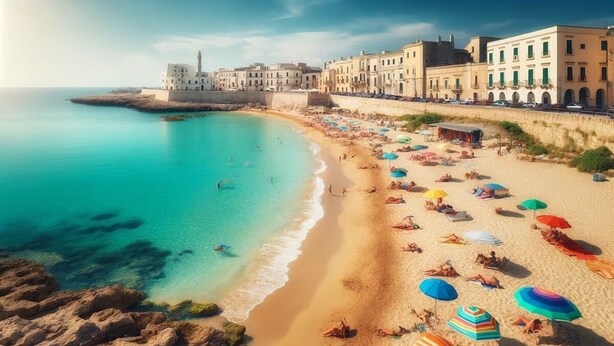 Una playa costera con un complejo de edificios históricos, aguas turquesas cristalinas y numerosos sombrillas de colores en la arena.