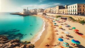 Ein Küstenstrand mit einem historischen Gebäudekomplex, klarem türkisblauem Wasser und zahlreichen bunten Sonnenschirmen im Sand.