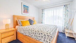 A neatly arranged bedroom with a wooden bed, a bedside table with a lamp, a framed artwork on the wall, and a window with sheer curtains. - Costas Smerelda 8, 16 Soldiers Point Rd - Linen, Views, Air Con, Wi-Fi (Soldiers Point)