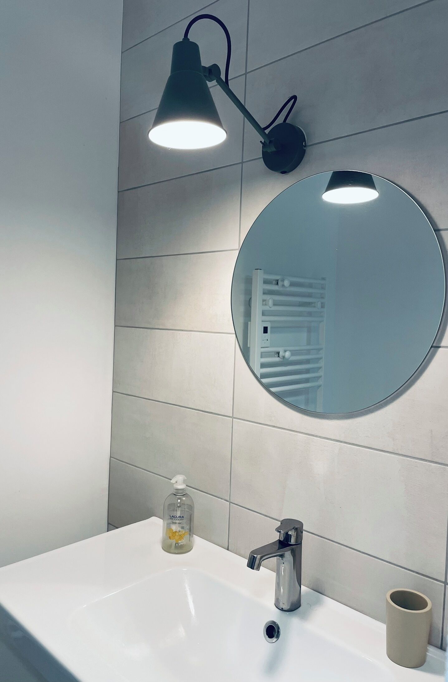A bathroom with a white sink, a round mirror, a black wall-mounted lamp, and a beige cup.