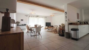 A spacious living area with a dining table, chairs, a sofa, a TV, and a kitchen counter.