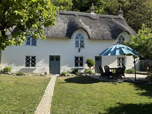 Ein Haus mit einem Strohdach, einem Garten, einem gepflasterten Weg und einem Tisch mit Stühlen unter einem blauen Sonnenschirm.