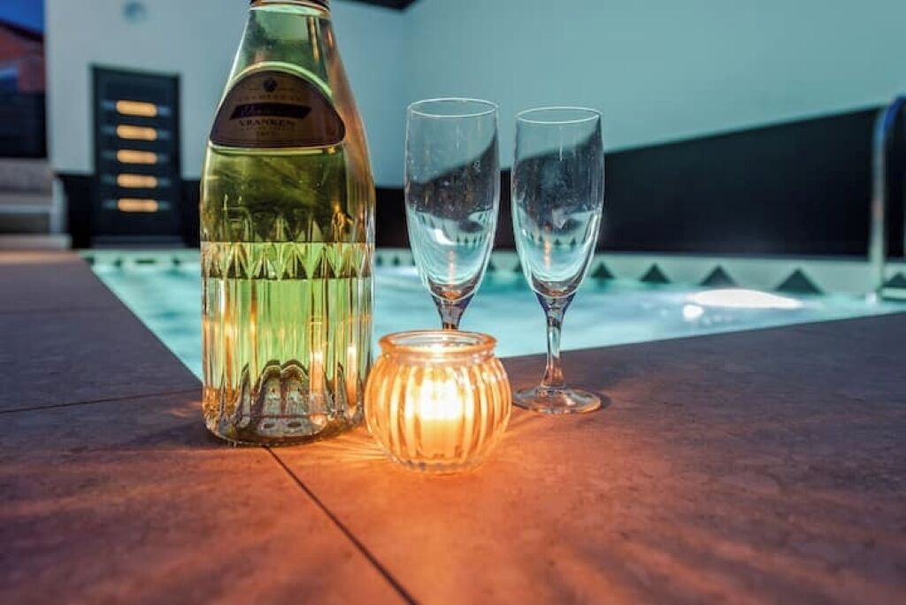 A bottle of wine, two glasses, and a candle on a table by a pool.