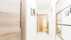 Ein Flur mit Holzdielen, weißen Wänden und gerahmten Bildern. Eine Tür führt in einen weiteren Raum, in dem sich eine Pflanze und ein kleiner Tisch befinden.