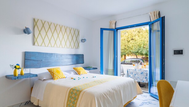 Ein Schlafzimmer mit einem Bett, einem fensterartigen Anblick in blauem Rahmen, einem gelben Stuhl und einem Tisch mit einer blau-weißen Tischdecke.
