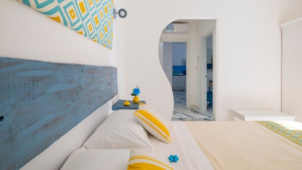 Une chambre à coucher avec une tête de lit en bois, un lit recouvert d’une literie jaune et blanche, une petite table sur laquelle est posé un vase, et une vue sur une autre pièce, accessible par une arche.