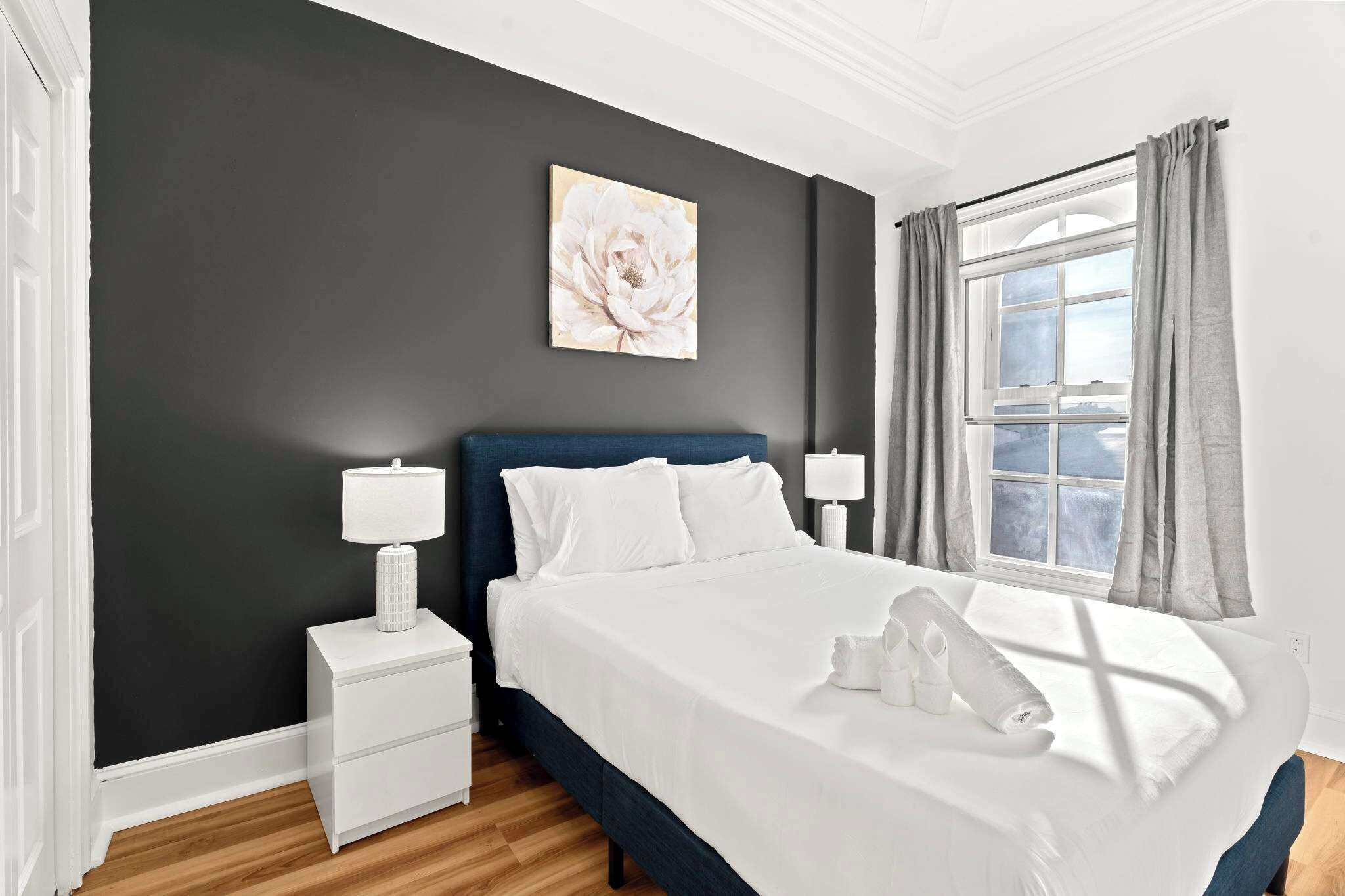 A hotel room with a bed, two bedside tables, a window with curtains, and a painting on the wall.