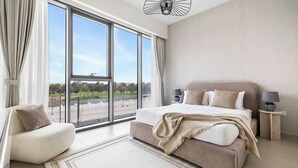 Et moderne soveværelse med et stort vindue, en seng med beige sengegavl, et natbord med en lampe og en stol.