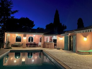 Un espace piscine avec une terrasse carrelée, une table à manger avec des chaises et un coin bar avec un comptoir et des tabourets.