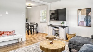 Ein modernes Wohnzimmer mit Sofa, Hocker und rundem Couchtisch. Es gibt einen Essbereich mit Tisch und Stühlen sowie eine Küchenzeile mit Fernseher und Schränken.