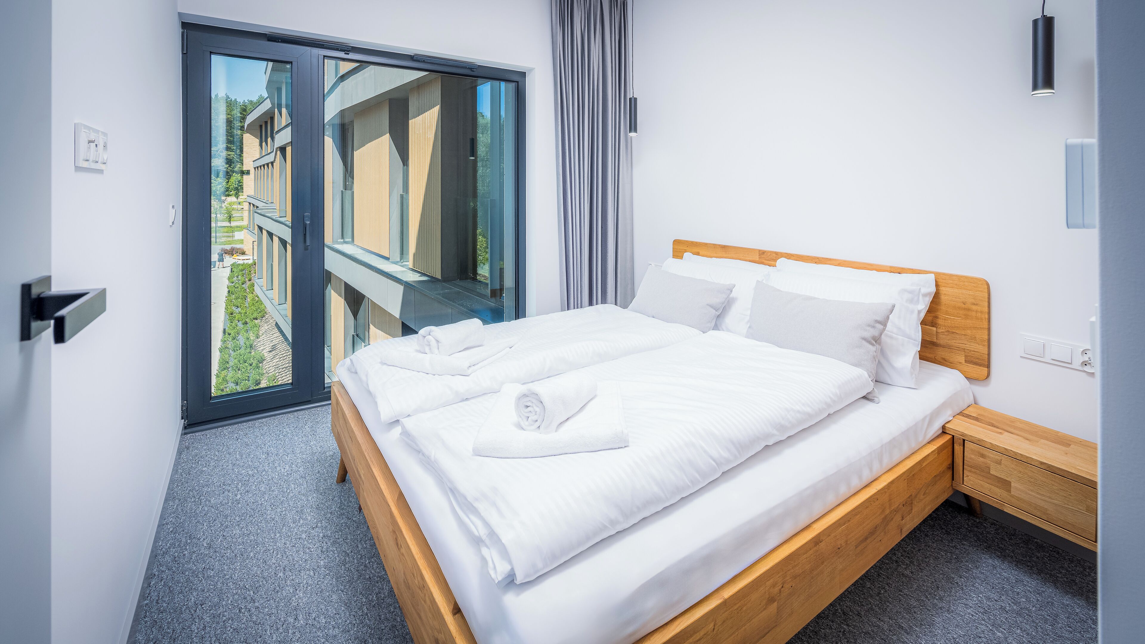 A modern hotel room with a large bed, a wooden headboard, a small bedside table, a nightstand, a sliding glass door to an outdoor area, and a view of the outside.